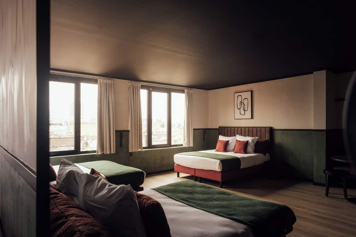 Ein Hotelzimmer mit einem Bett, zwei Nachttischen, einem Sessel und einem Fenster mit Vorhängen, mit Blick auf die Stadt.