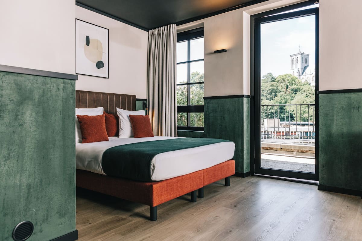 Ein modernes Hotelzimmer mit einem großen Bett, Blick auf eine Stadtlandschaft durch ein Fenster und einer grünen Akzentwand.
