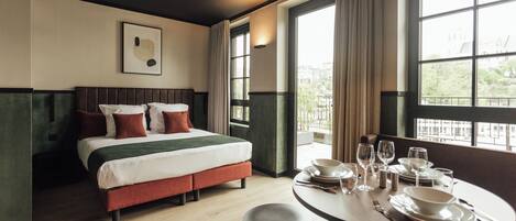 A hotel room with a bed, a dining table set for two, and large windows with curtains.