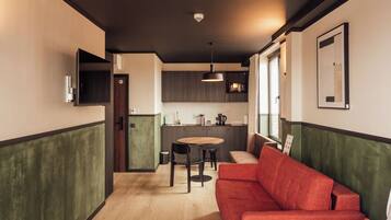 A compact hotel room with a bed, a red sofa, a dining table with chairs, a kitchen area, and a large window.