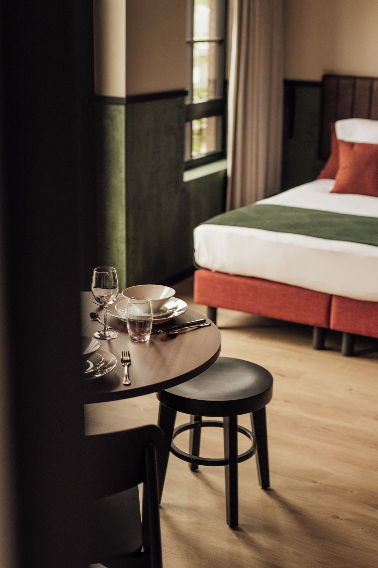 Ein ordentlich gedeckter Esstisch mit Stuhl, ein Bett mit rotem Kopfkissen und ein Fenster mit Vorhängen in einem Hotelzimmer.