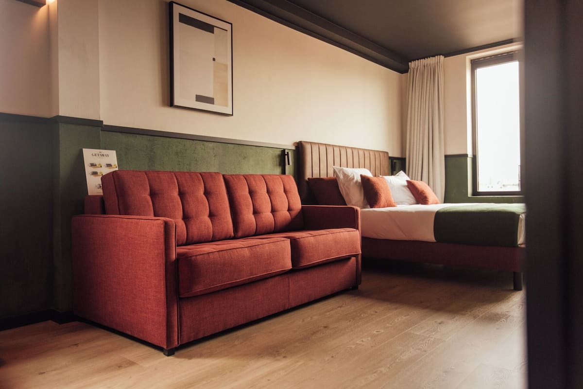 Ein Zimmer mit einer roten Couch, einem Bett mit weißen und orangefarbenen Kissen, einem gerahmten Bild an der Wand und einem Fenster mit Vorhängen.