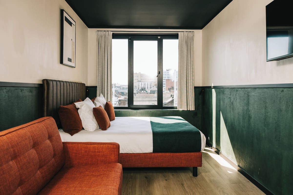 Ein Hotelzimmer mit einem Bett, einem roten Sofa, einem großen Fenster mit Vorhängen und einem gerahmten Bild an der Wand.