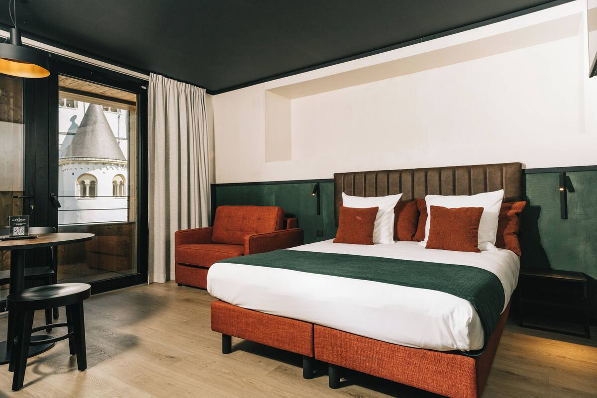 Ein Hotelzimmer mit einem großen Bett, einer Sitzecke mit Tisch und Stühlen und Blick auf ein Gebäude durch ein Fenster.