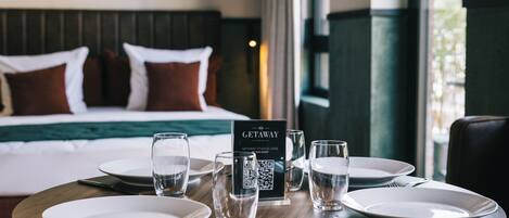 Una habitación de hotel con una mesa de comedor bien puesta, una cama con edredón verde y un cuadro en la pared.