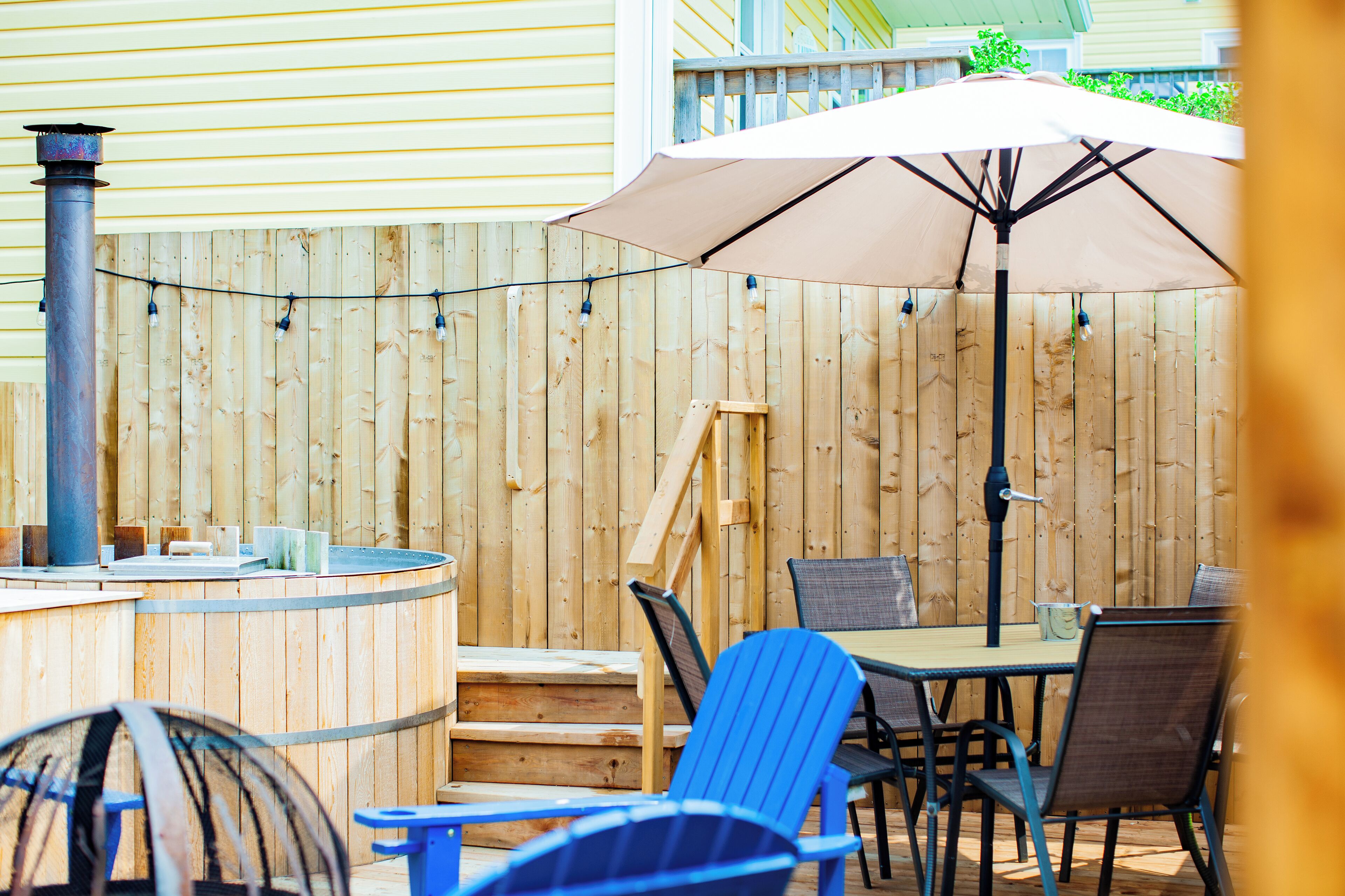 Une terrasse en bois avec un jacuzzi, une table entourée de chaises et un parasol.