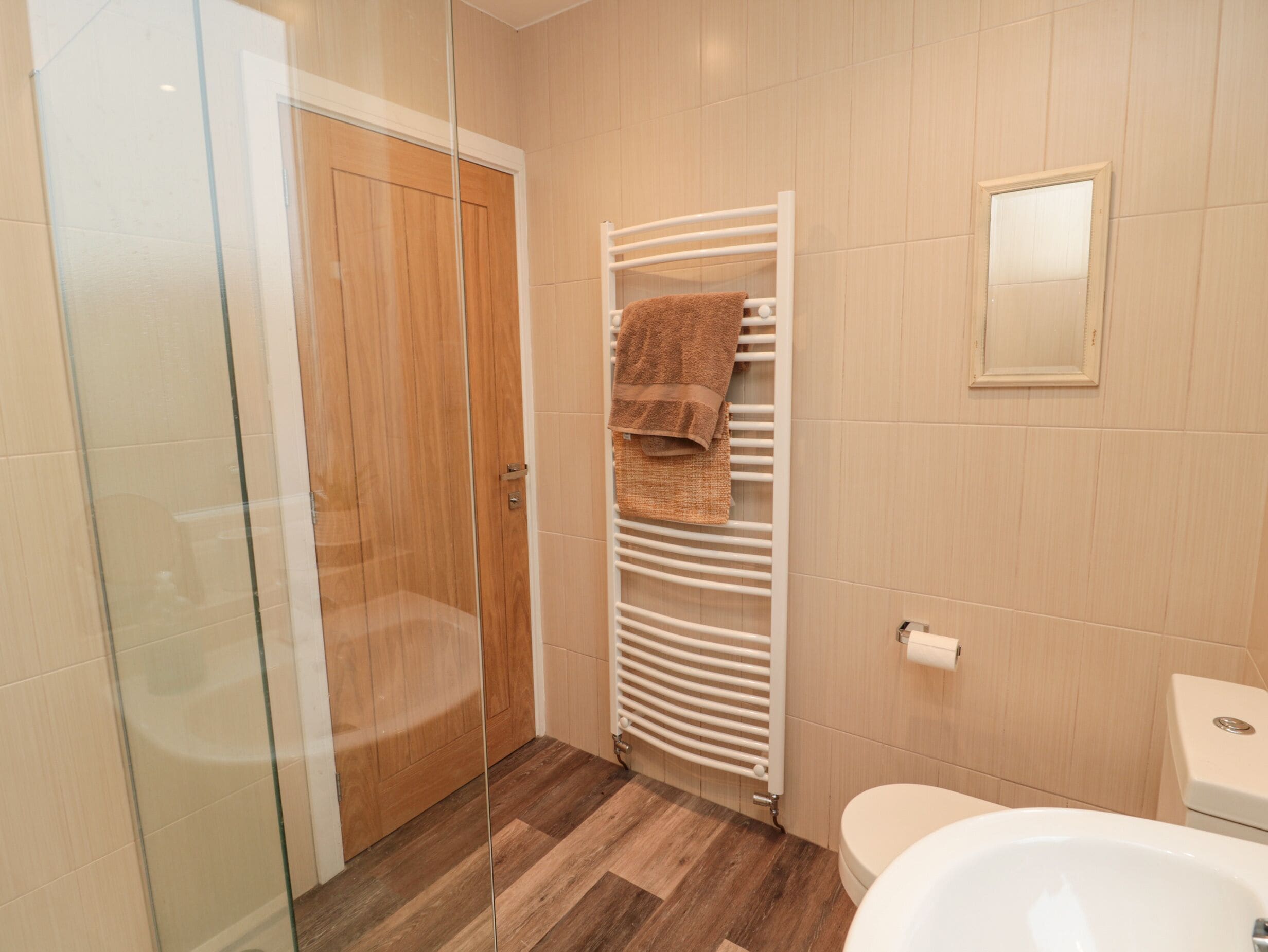 Casa de banho com porta de madeira, cabine de duche em vidro, um porta-toalhas com uma toalha castanha, radiador branco e lavatório branco.