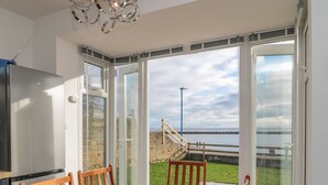A dining area with a table and chairs, a glass-top table, and a view of a coastal landscape through large windows.