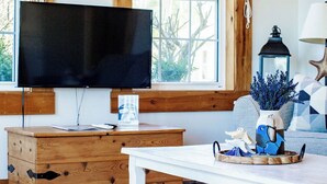Une pièce avec un plancher en bois, une table blanche, un téléviseur posé sur un meuble en bois et une fenêtre donnant sur des arbres.