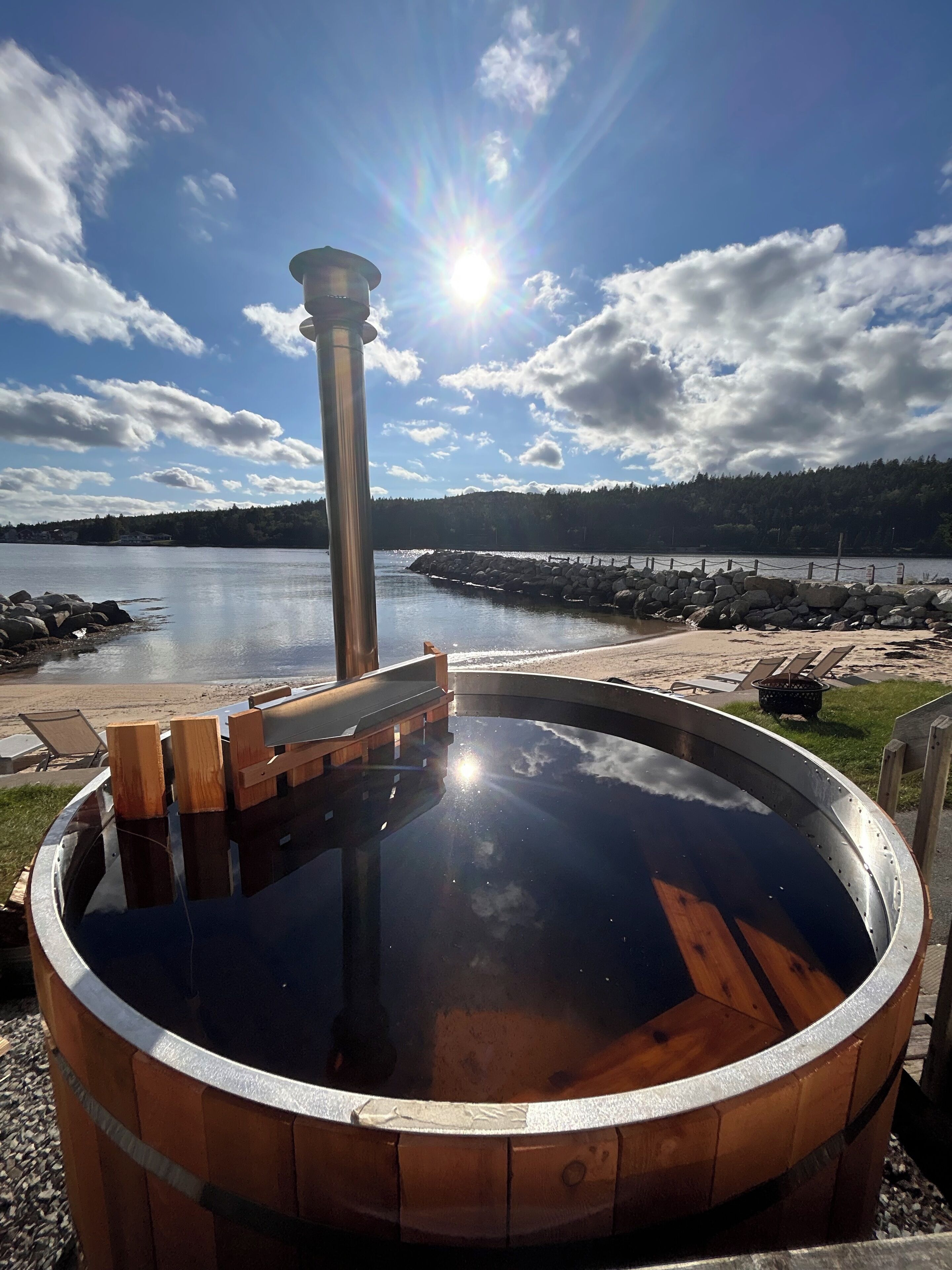 Un bain à remous doté d’une terrasse en bois, entouré d’un mur de pierres, offrant une vue sur un lac aux rives rocheuses et sur une colline boisée en arrière-plan.