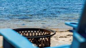Une chaise de plage et un foyer au bord d’une plage de sable, avec vue sur un lac paisible et une colline boisée en arrière-plan.