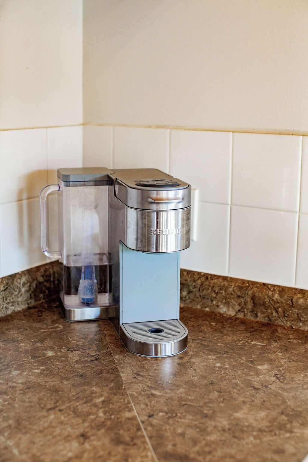 Una cafetera con depósito de agua transparente y base metálica sobre una encimera de cocina.