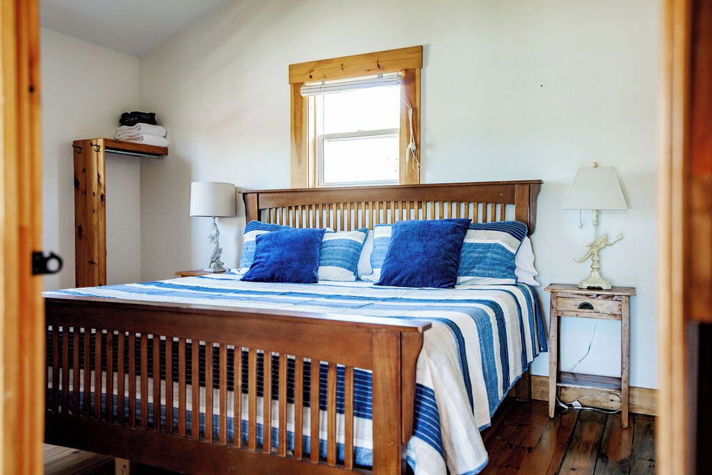 Een slaapkamer met een houten bed, gestreept beddengoed, een nachtkastje met een lamp en een raam boven het bed.