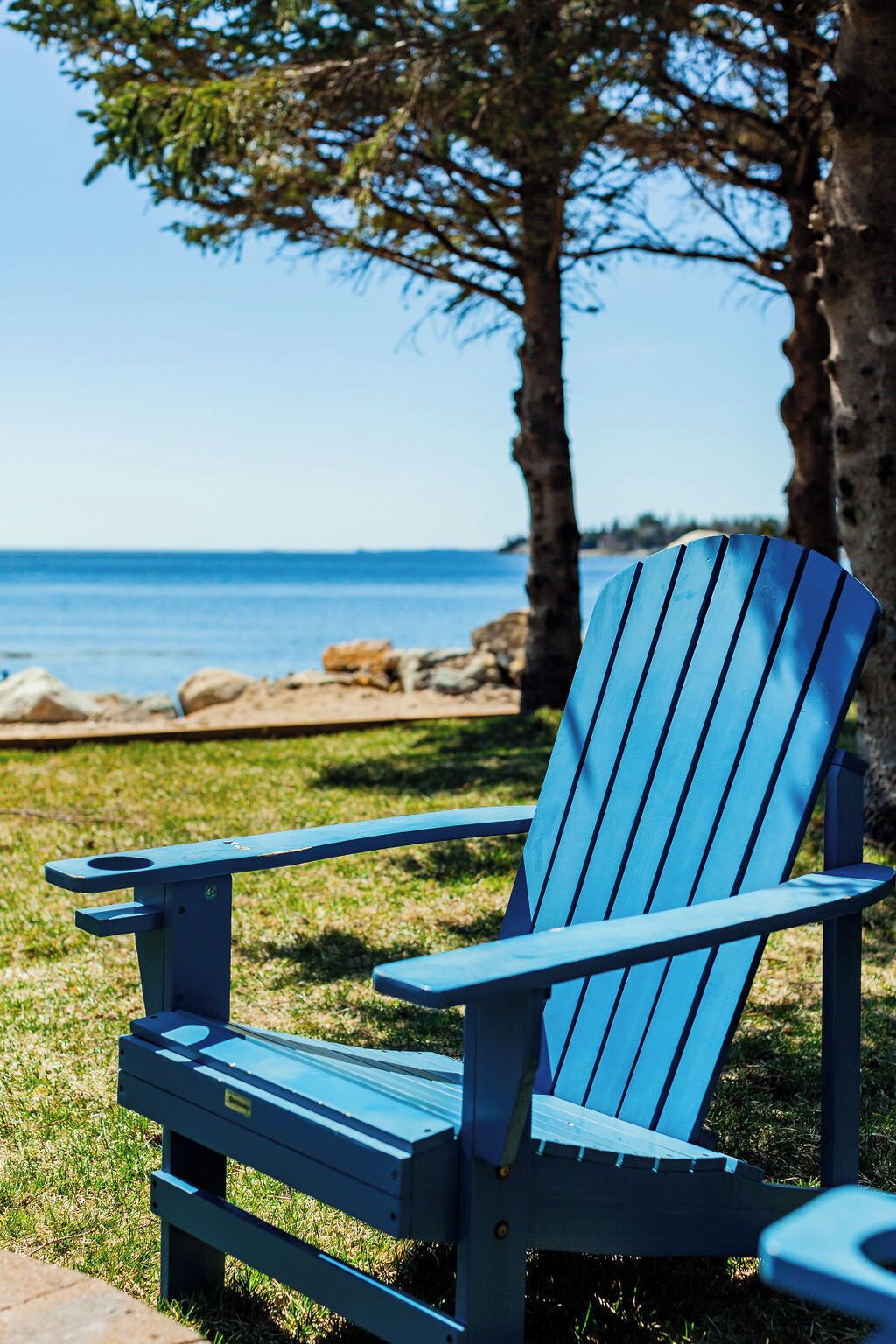 Uma cadeira Adirondack azul com estrutura de metal, posicionada sobre a grama, com vista para o oceano e costa rochosa ao fundo.