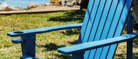 Uma cadeira Adirondack azul com estrutura de metal, posicionada sobre a grama, com vista para o oceano e costa rochosa ao fundo.