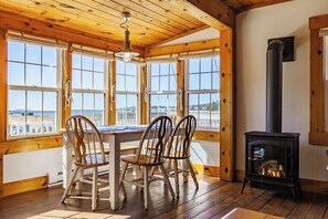 A cozy room with a wooden table and chairs, large windows, and a wood-burning stove. - Oceanfront 2-Bedroom Cottage | Private Hot Tub | Wood Stove | Near Halifax (Boutiliers Point)