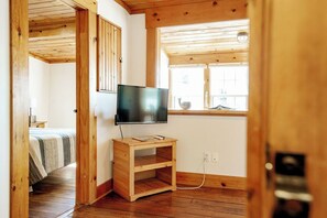 A room with a wooden ceiling, a television on a wooden stand, a window with a view, and a bed with a striped blanket. - Oceanfront 2-Bedroom Cottage | Private Hot Tub | Wood Stove | Near Halifax (Boutiliers Point)