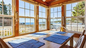 Un coin repas baigné de lumière naturelle, avec une table et des chaises en bois, de grandes fenêtres offrant une vue sur la mer et un plafond en bois.
