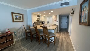 Une cuisine dotée d’un îlot central, de chaises en bois et d’une table à manger. On y trouve un plan de travail avec des appareils électroménagers et un évier. Les murs de la pièce sont peints en bleu clair et le sol est recouvert de parquet.