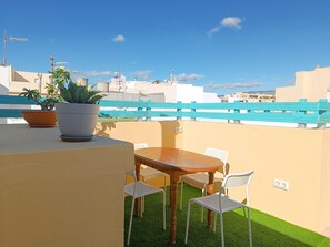 Terraza en la azotea con mesa de madera y sillas blancas, rodeada de plantas en macetas.