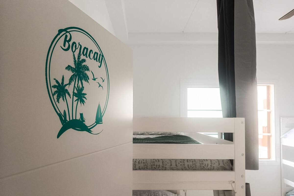 A bunk bed room with a wall decal featuring palm trees and birds, a window with curtains, and a view of the outside.