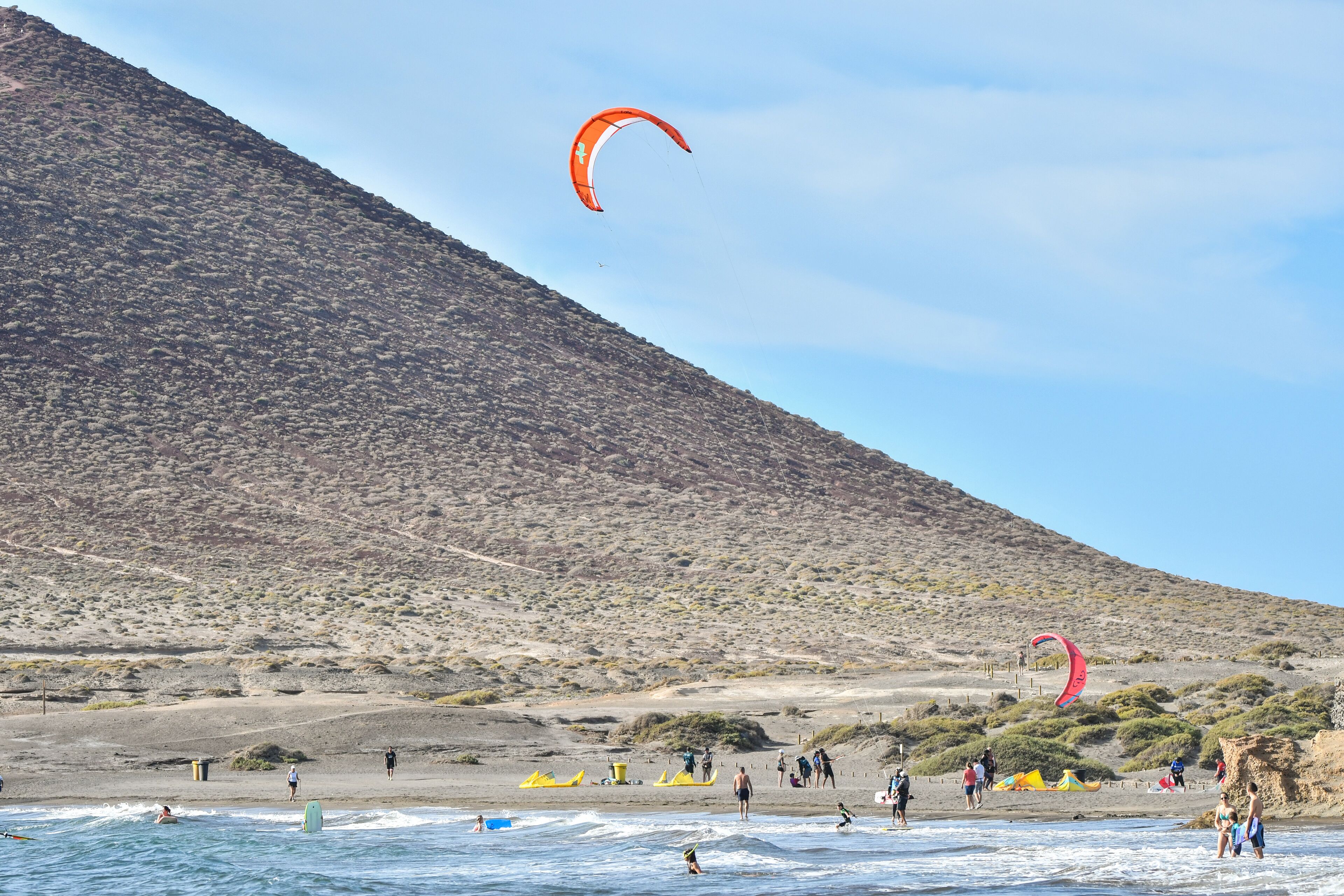 Uma praia com pessoas praticando atividades aquáticas, uma colina ao fundo e céu claro.