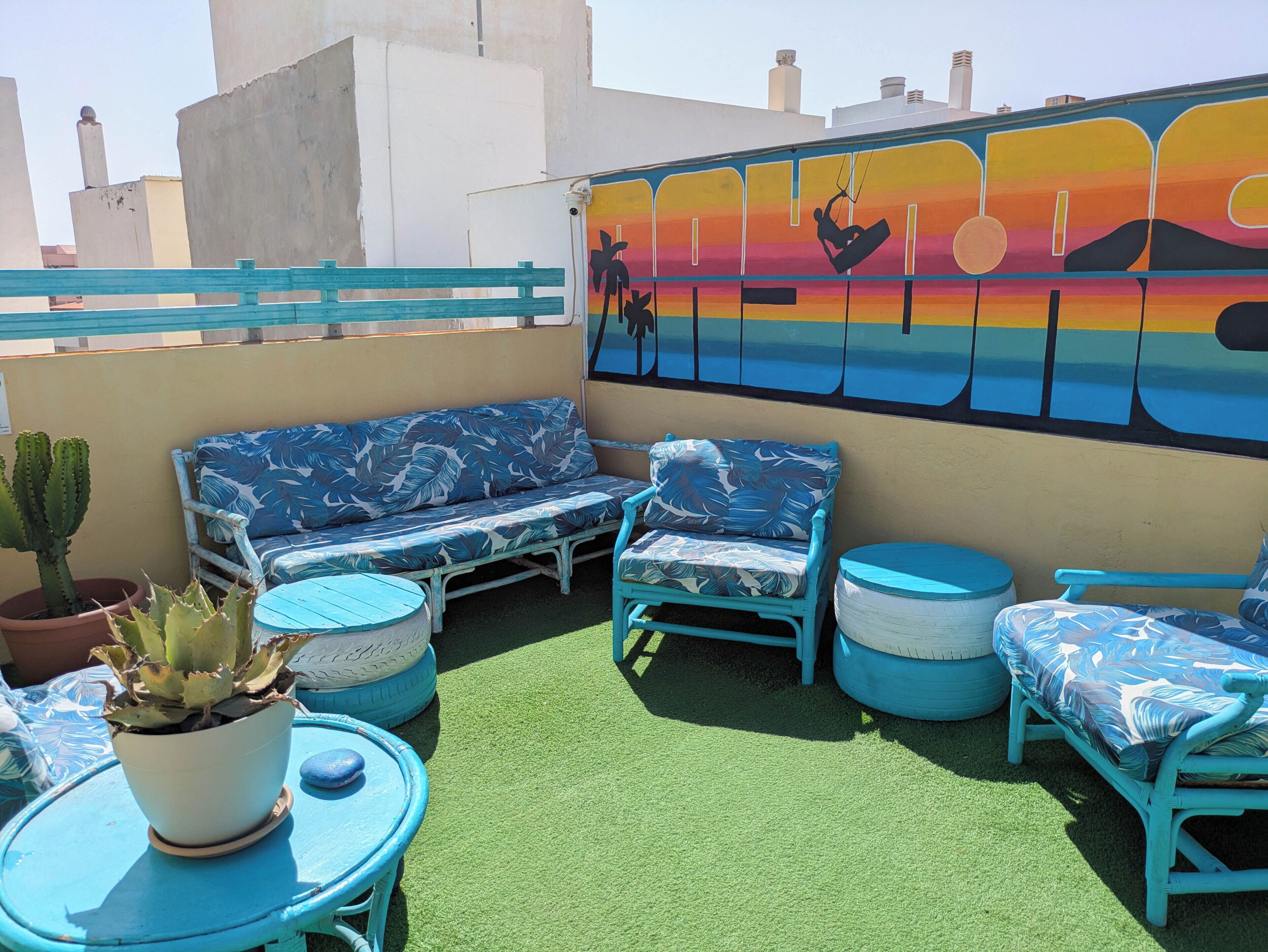 A rooftop terrace with blue furniture, a cactus plant, and a mural depicting a sunset scene.
