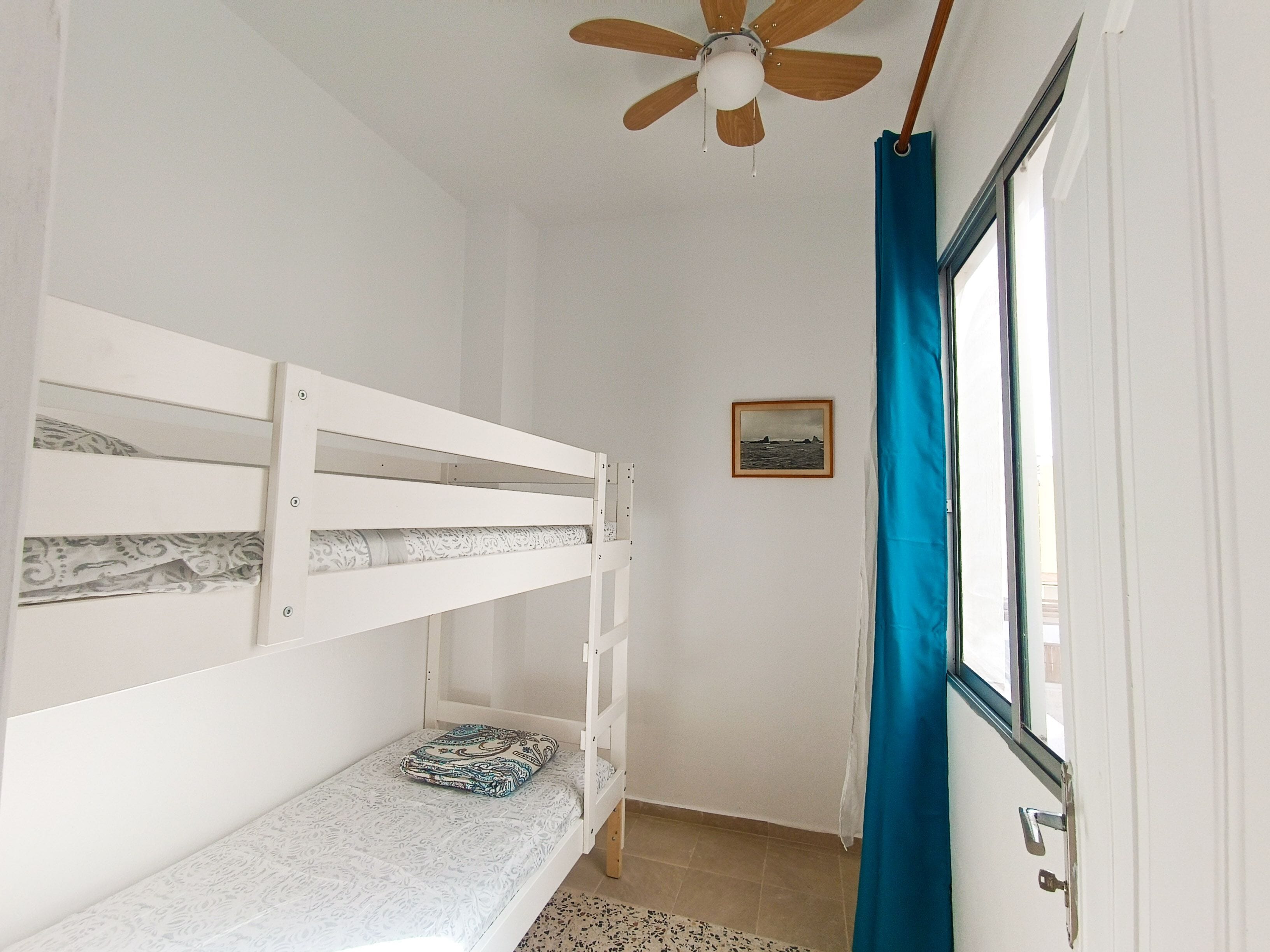A room with a bunk bed, a ceiling fan, a window with blue curtains, and a framed picture on the wall.