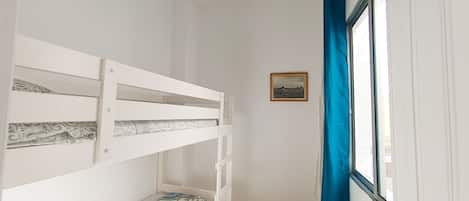 A room with a bunk bed, a ceiling fan, a window with blue curtains, and a framed picture on the wall.