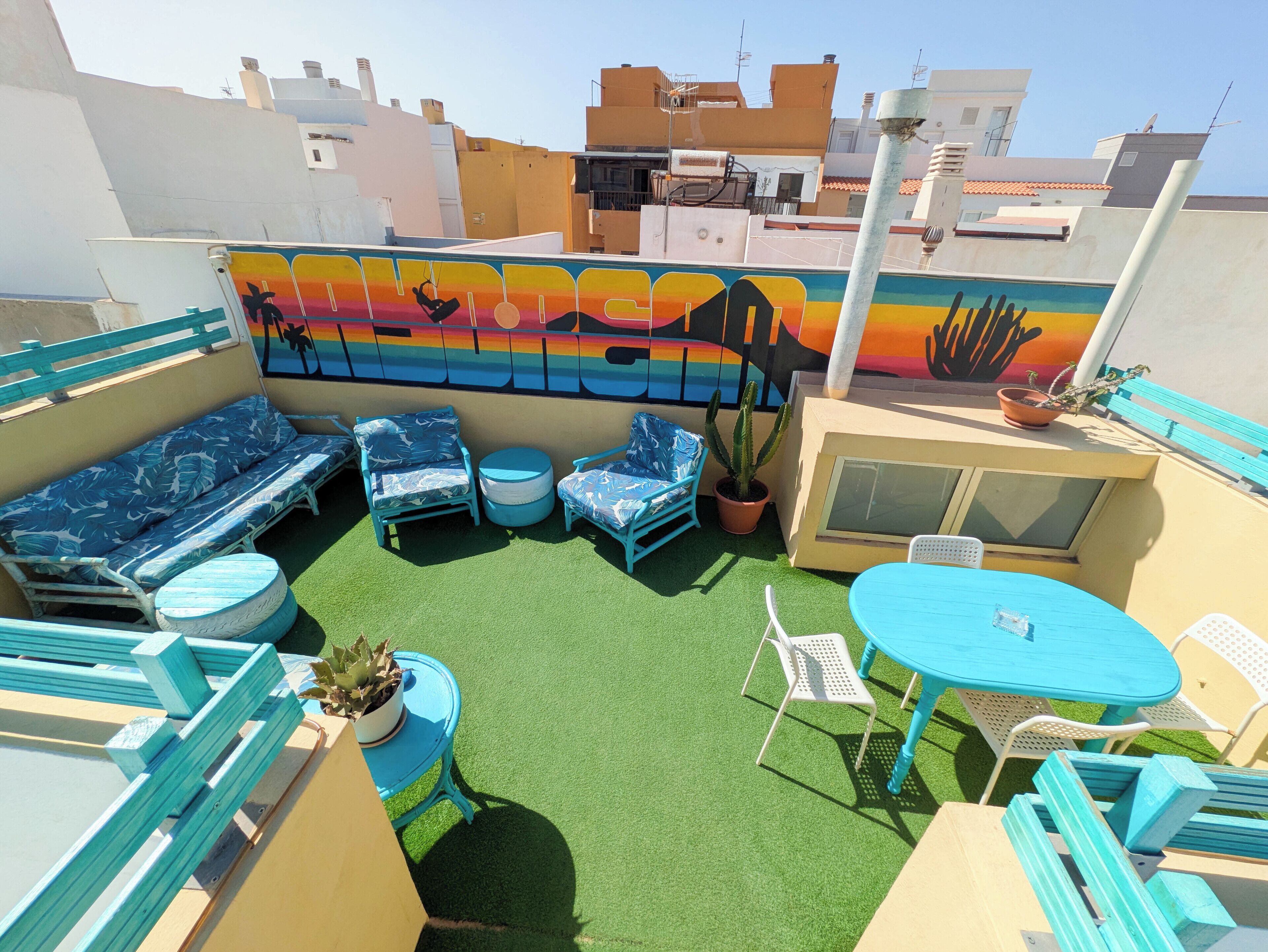 Un espace extérieur aménagé avec des sièges, une fresque murale, une pelouse artificielle verte et du mobilier bleu.