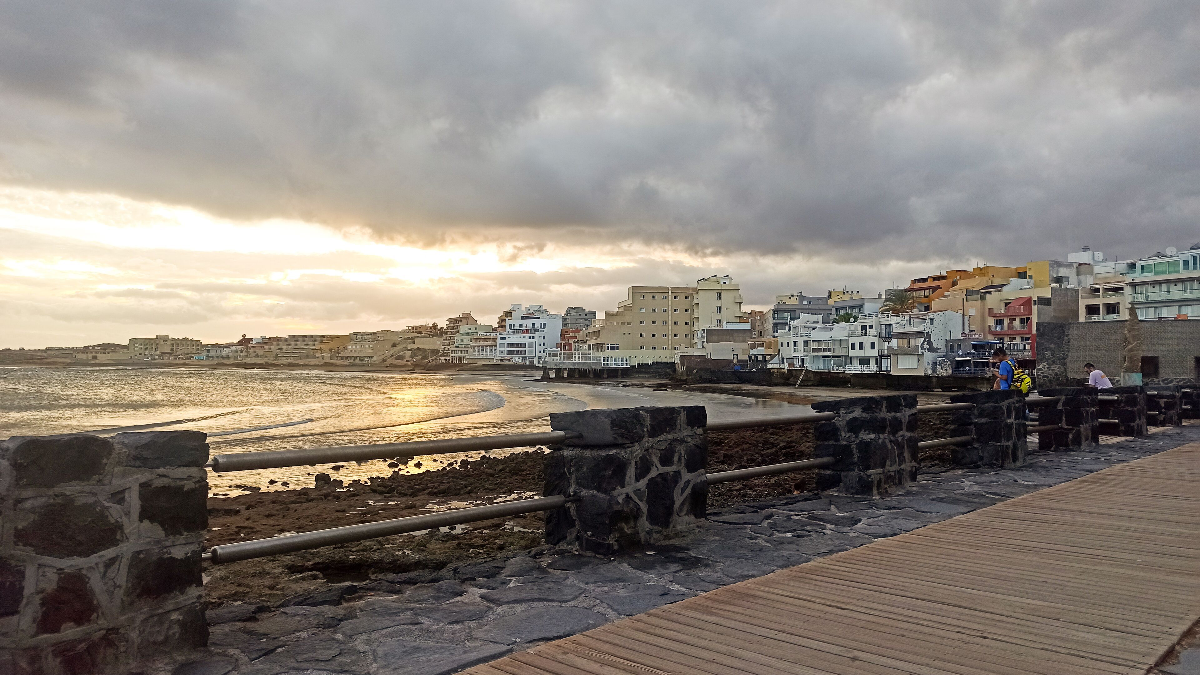 A coastal walkway with stone railings and a wooden deck, leading to a beach with buildings in the background.