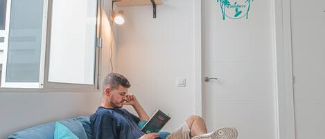 Eine Person sitzt auf einem blauen Sofa und liest ein Buch, darüber eine Holzregal mit Büchern und eine Wandsticker mit Palmen.