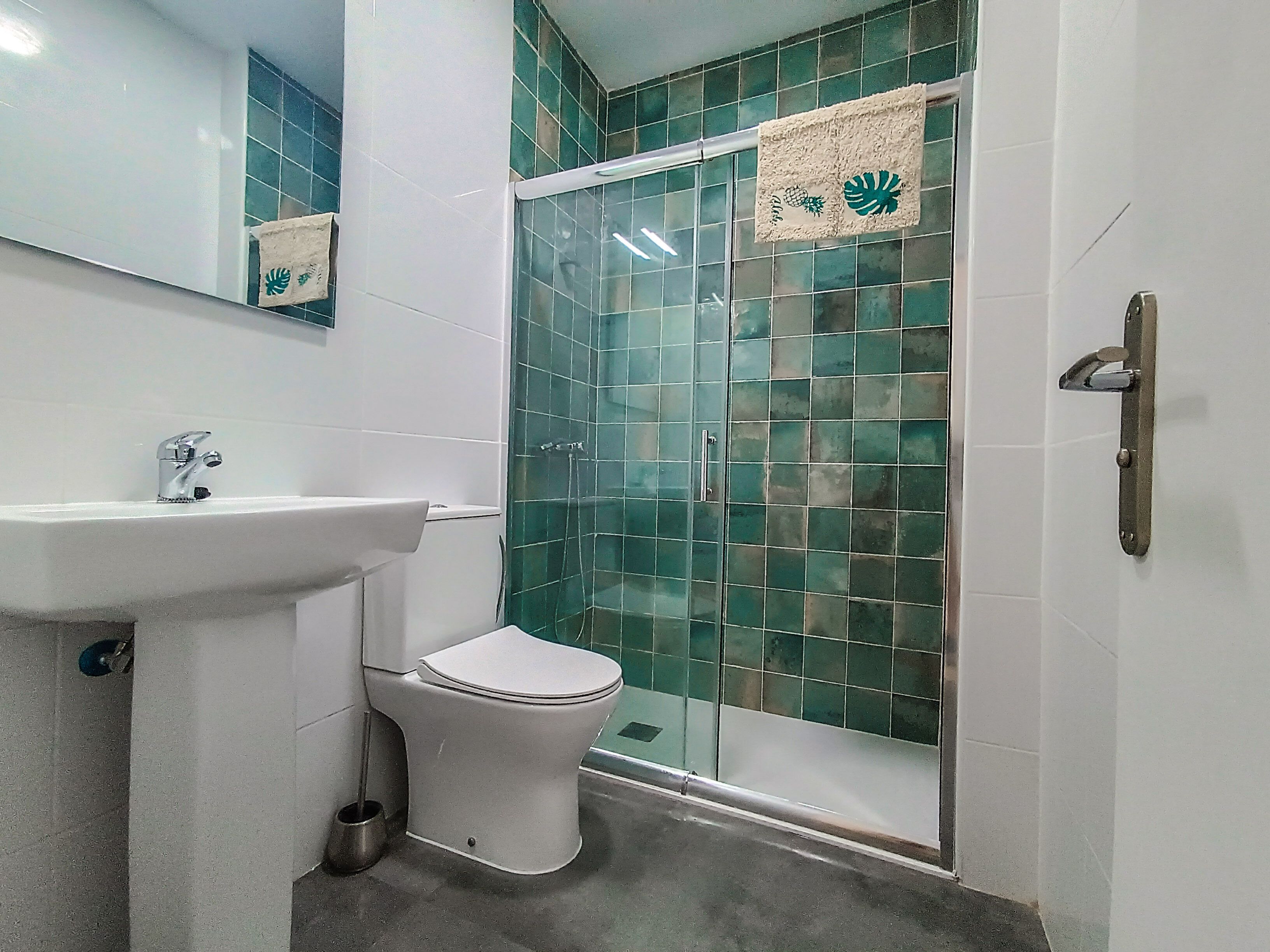 Une salle de bain avec un lavabo blanc, des toilettes et une douche en verre, dont les murs sont recouverts de carreaux verts.