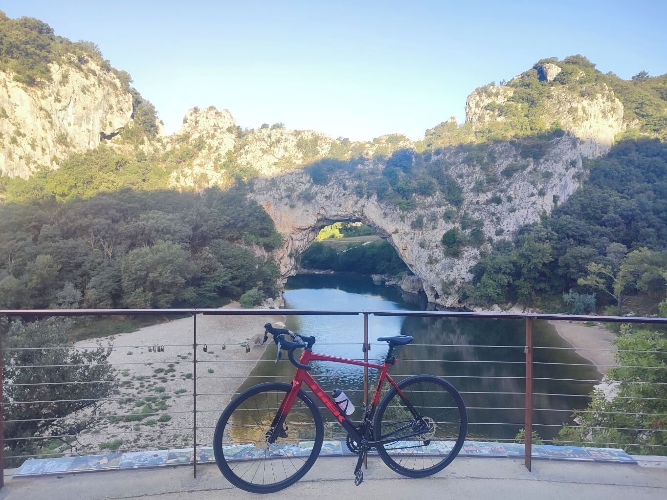 A red bicycle parked on a balcony overlooking a river with a natural rock bridge.