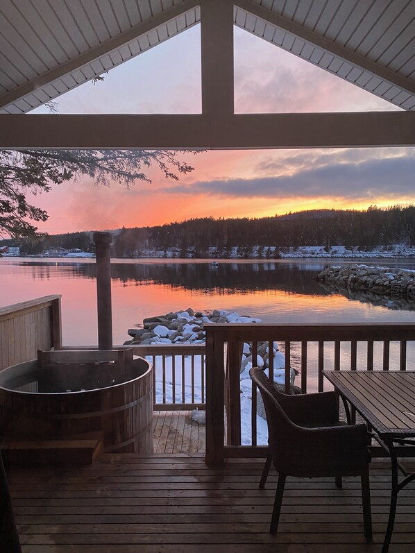 Una terraza de madera con un jacuzzi, una mesa y sillas, con vista a un lago tranquilo y una orilla boscosa al atardecer.