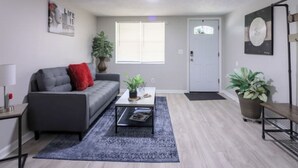 Un salon moderne avec un canapé gris, une table basse et un tapis bleu. On y trouve deux plantes en pot, une porte et un mur orné d’une œuvre d’art circulaire.
