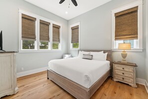 A bedroom with a large bed, a nightstand with a lamp, and three windows with blinds. - 30A Beach House - Beach Please (Santa Rosa Beach)