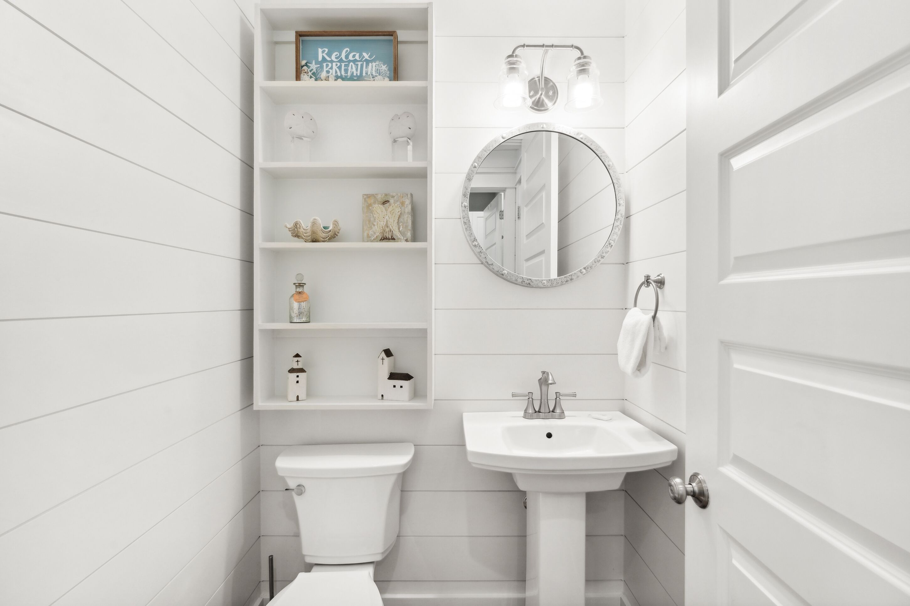 Une salle de bain blanche avec des toilettes, un lavabo et des étagères ornées d’objets décoratifs.