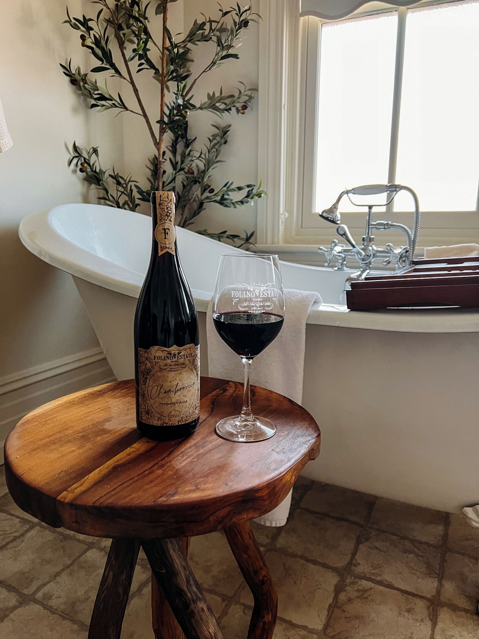 A bottle of Folino Estate champagne and a glass of red wine on a wooden stool in a bathroom with a bathtub and a window.