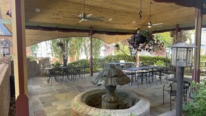 An outdoor patio with stone flooring, metal tables and chairs, a central stone fountain, hanging planters, and ceiling fans.