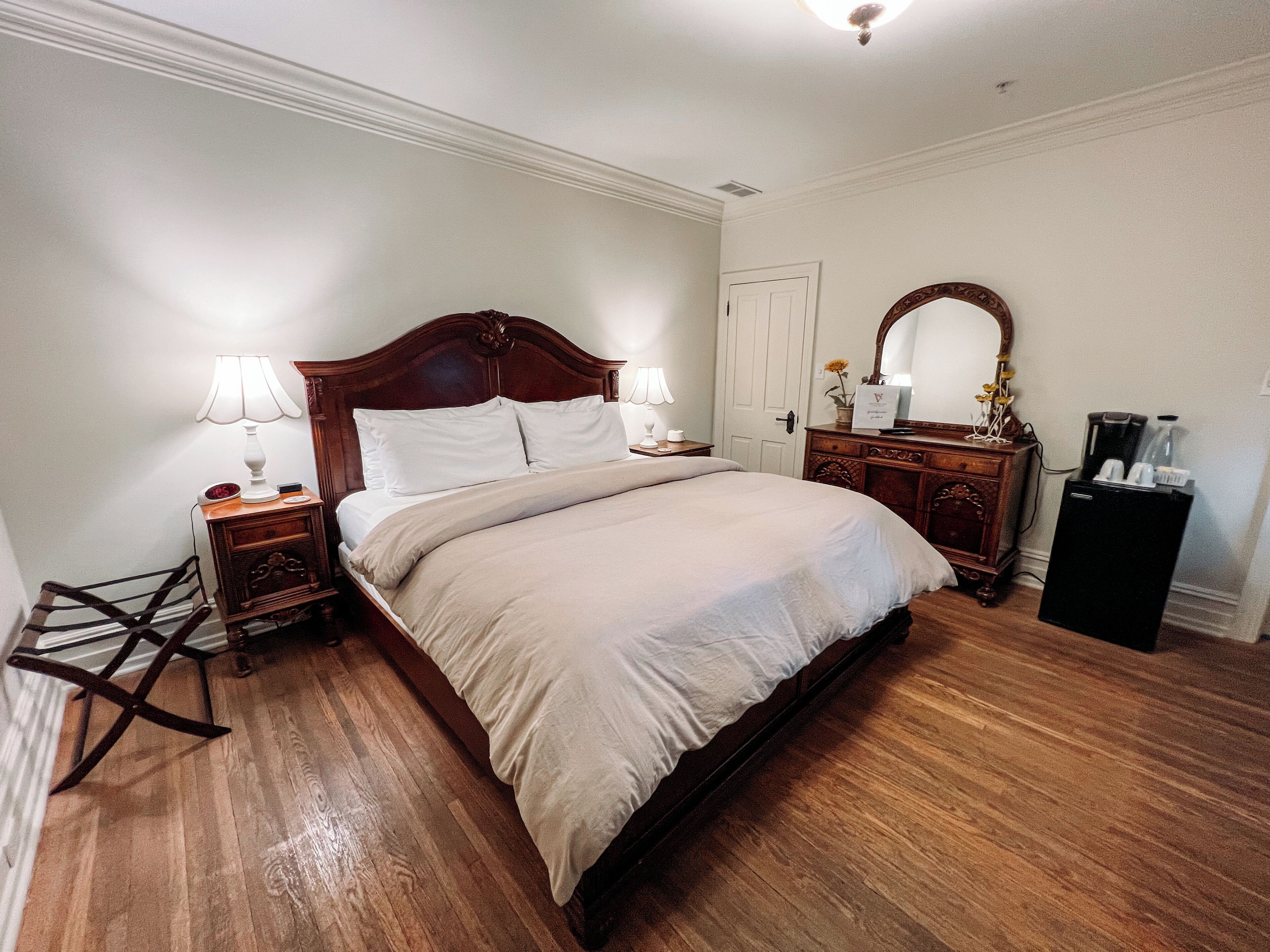 A neatly made bed in a room with wooden flooring, a dresser, a nightstand, and a small refrigerator.