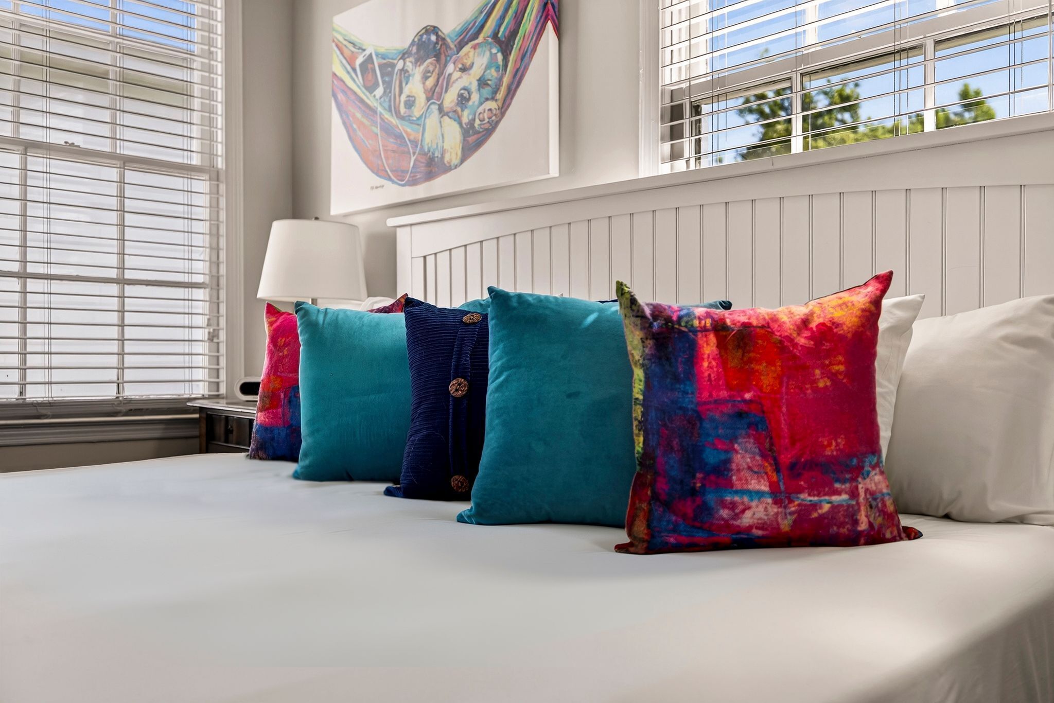 A bed with colorful pillows, a wall-mounted lamp, and a hammock painting.