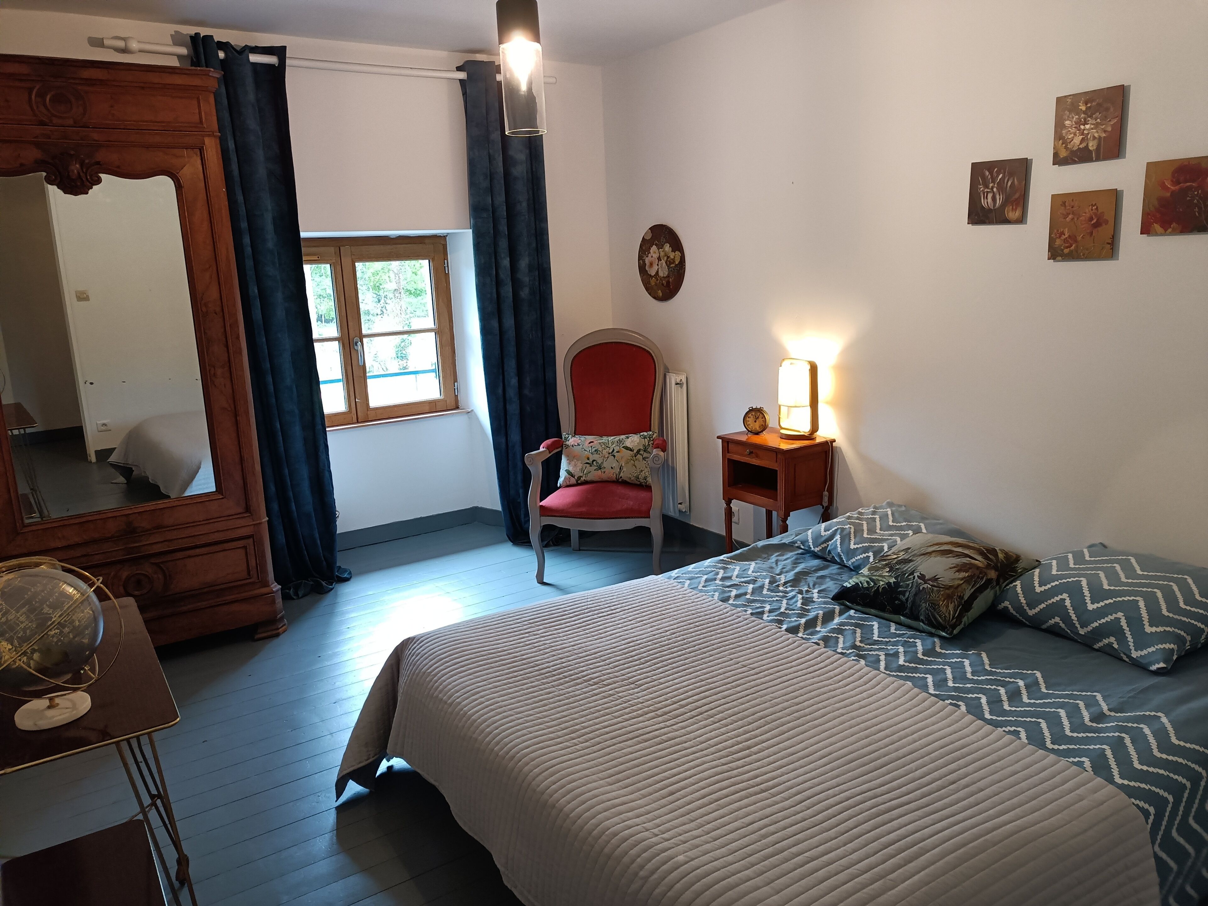 Un dormitorio con cama, una silla, una mesita de noche con lámpara, un espejo y una ventana con cortinas.