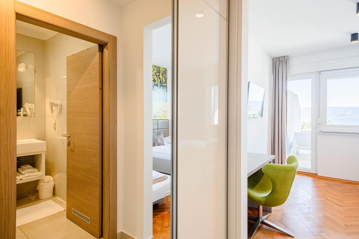 Quarto de hotel moderno com porta de madeira, banheiro com pia e suporte para toalhas, cama com cabeceira e área de refeições com mesa e cadeiras verdes.