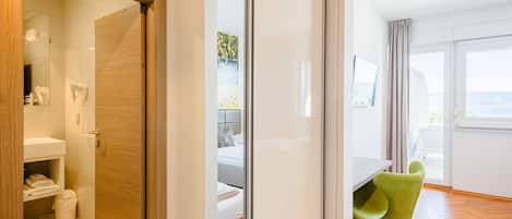 A modern hotel room with a wooden door, a bathroom with a sink and towel rack, a bed with a headboard, and a dining area with a table and green chairs.