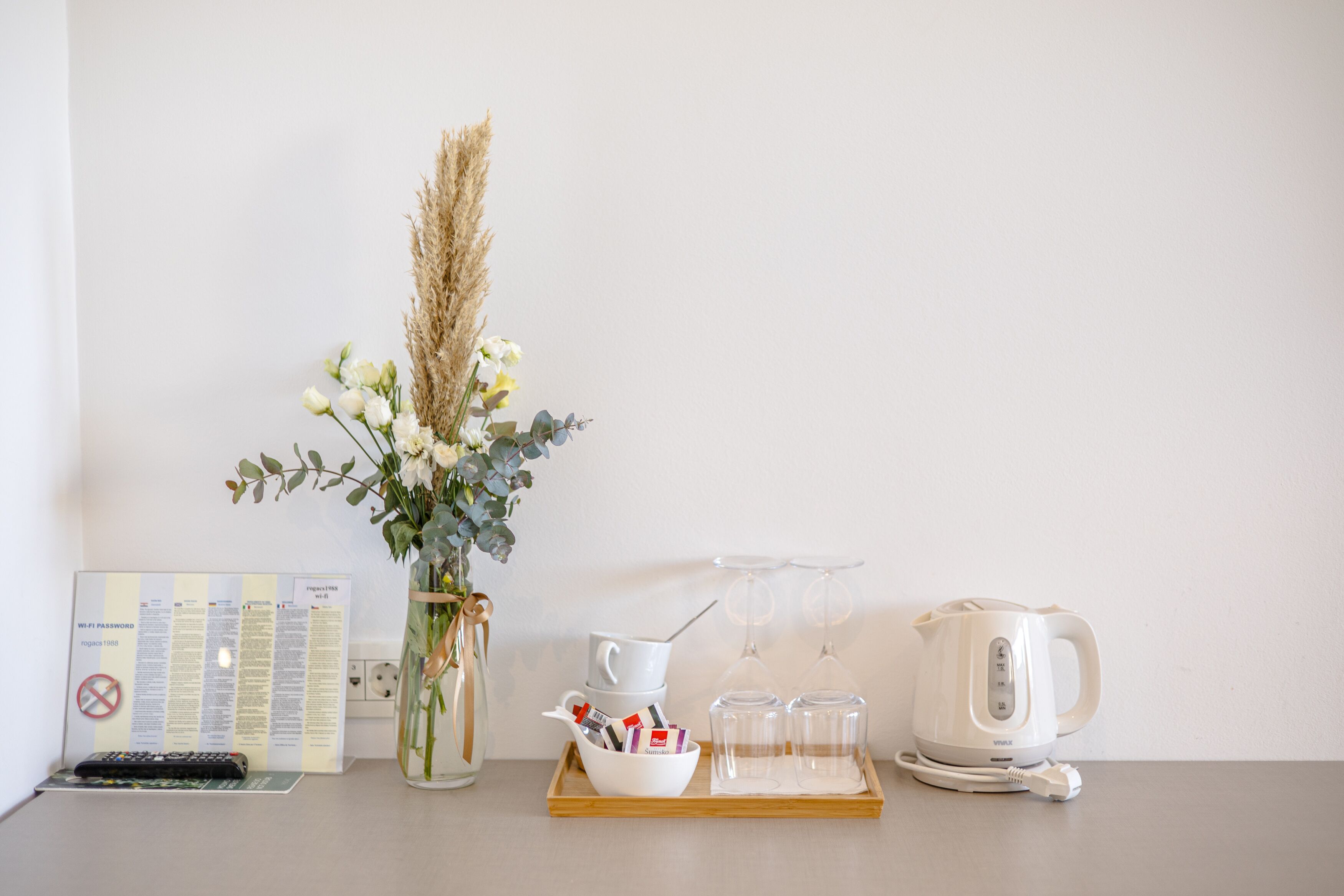 A clean, minimalist table with a vase of flowers, a tray with a kettle, cups, and a remote control.