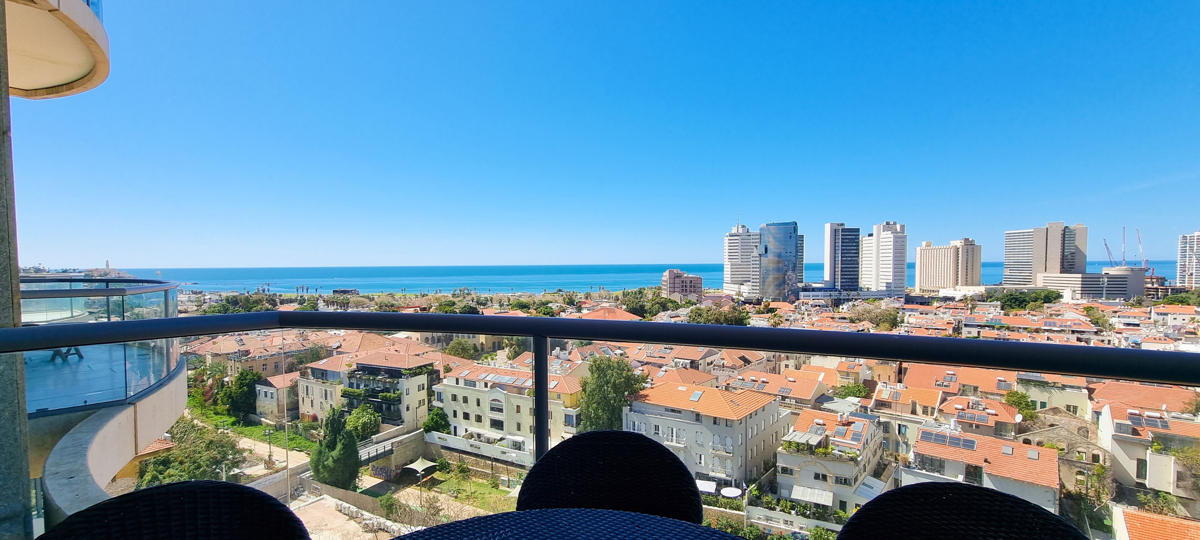 Un balcon offrant une vue sur un paysage urbain côtier, avec des bâtiments et un ciel bleu dégagé.