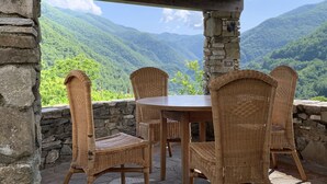 Ein überdachter Außenbereich mit Korbstühlen und einem Tisch bietet Blick auf eine gebirgige Landschaft.