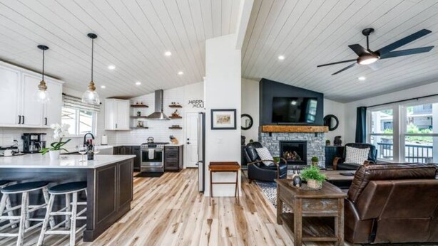 A modern kitchen with white cabinets, a central island with bar stools, a dining area with a fireplace, and a living room with a brown leather sofa and a ceiling fan. - Stunning New Horse and Hare Inn (Murphys)