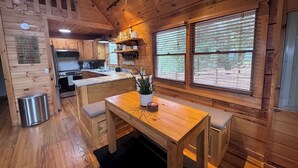 Ein Interieur einer Holzhütte mit Holzwänden, einer Küchenzeile, einem Esstisch und einem großen Fenster mit Jalousien.